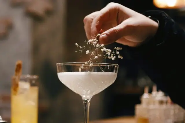 A hand decorating a margarita glass filled with a white opaque liquid, with a branch of a white plant. Surrounding it is a cosy cafe or bar setting.