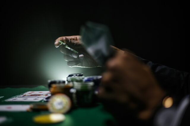 Hand holding poker chips over piles of poker chips and playing cards sitting on the table