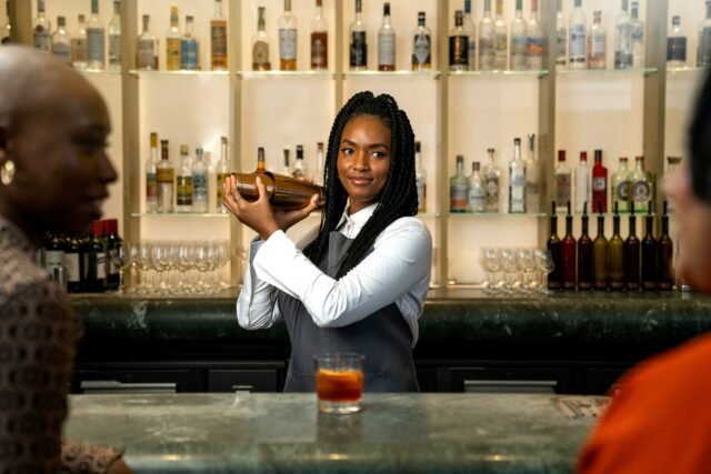 A bartender standing behind at a bar shaking a cocktail shaker preparing a drink in a brightly lit bar or restaurant
